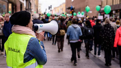 Demonstranter går på Alexandersgatan i Helsingfors. Kvinna talar i en megafon.