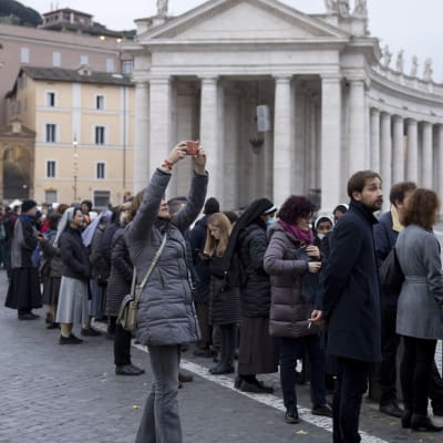 Människor som köar med kolonnaden vid Petersplatsen i bakgrunden. Bland de köande syns flera ordenssystrar..