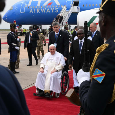 Påve Franciskus i rullstol på röda mattan i Kinshasa. Han mottas av ett hedersgarde av kongolesiska soldater.