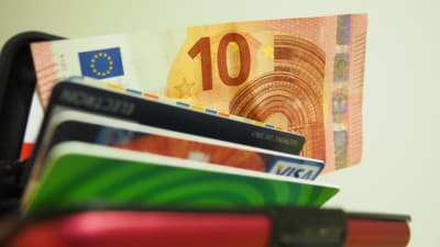 En tioeurossedel kommer fram ur en plånbok. Ett busskort och bankkort syns också delvis.