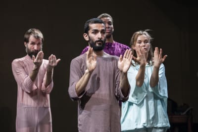 Fyra dansare i pastellfärgade kläder på en mörk scen håller upp händerna framför sig. 