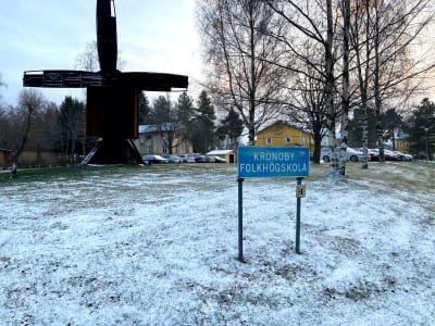Skylt med texten "Kronoby folkhögskola". I bakgrunden står en kvarn.