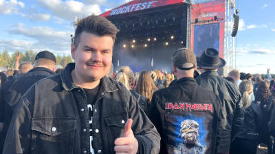 En man håller uppe tummen på rockfestival med scen i bakgrunden.