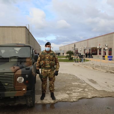 Sotilas seisoo maastokuvioisessa univormussa maastokuvioiden jeepin vieressä. Takana näkyy kaksi betoninharmaata rakennusta.
