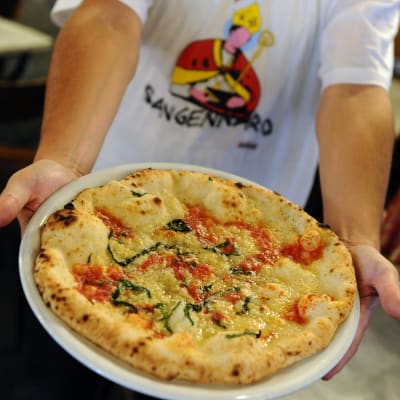 Mies ojentaa lautasta, jolla pizza, vieressään Napolin suojeluspyhimystä San Gennaroa esittävä pysti.