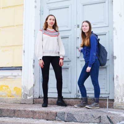 Två unga kvinnor står på trappan till Lovisa Gymnasium, ett gult trähus med blå ytterdörr. Den ena av dem håller i dörrhandtaget.