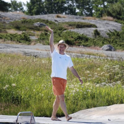 Riko Eklundh går på en brygga och vinkar. Han har orangerutiga shorts, vit t-skjorta och en halmhatt på sig.