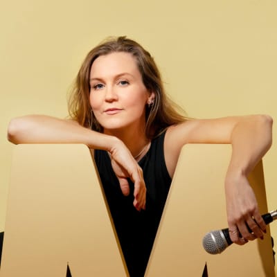 Märta Westerlund framför en enfärgad bakgrund lutar mot ett stort M och håller i en mikrofon.