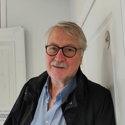En äldre man står utanför en dörröppning. Inne i rummet syns färggranna tavlor på väggarna.