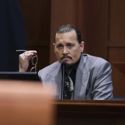 Johnny Depp håller i ett par solglasögon medan han sitter i ett vittnesbås. Framför honom en mikrofon.