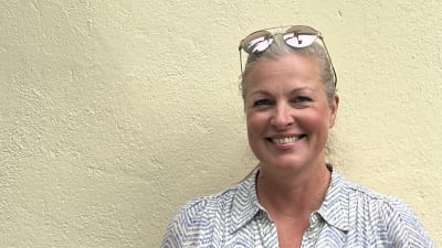 En person med blont bakåtkammat hår, solglasögon i pannan och en blåvit mönstrad klänning tittar rakt in i kameran.