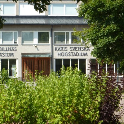 En byggnad skymtar bakom träd och buskar, det är Karis-Billnäs gymnasium.