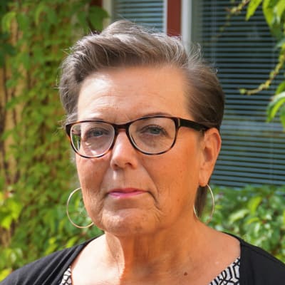 Kvinna med kort hår och glasögon står framför en gul byggnad.