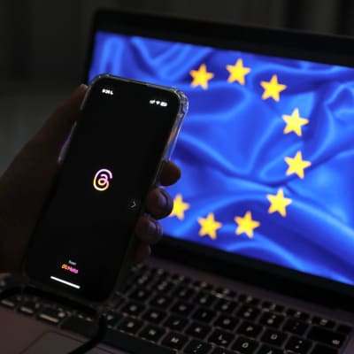 Smartelefon med Threads logga hålls upp framför en dator med EU:s flagga