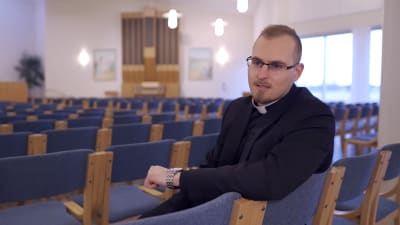 En präst sitter på en stol i en stor sal och pratar.