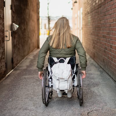 Rullstol bakifrån, en ung person med långt, blont hår. Röd tegelvägg på sidan. 