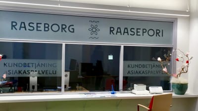 En kundbetjäning där personalen ska finnas bakom ett glas med texten Raseborg Raasepori på glaset. Stolar och litet datorbord syns framför disken. Inga människor på plats.
