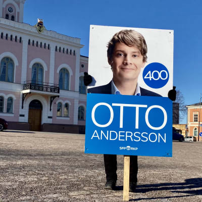 En person som inte syns håller upp en skylt med en valaffisch på Otto Andersson. Personen med skylten står på Lovisa torg och det rosa stadshuset syns i bakgrunden.
