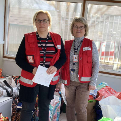 Kaksi naista, Tarja Kaskama ja Maria Pietikäinen, seisovat huoneessa, jossa on paljon kauppakasseja lattialla. Naisilla on SPR:n punaiset liivit yllään. 