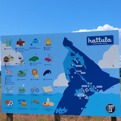 Sinisessä kyltissä on Hattulan kartta. Taustalla viljapeltoa ja taivaalla poutapilviä.
