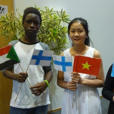Tre barn står med flaggor i hand. En sudansk pojke viftar med Sudans och Finlands flaggor, en vietnamesisk flicka med Vietnams och Finlands flaggor och en bosnisk flicka med Bosniens och Tysklands flaggor.