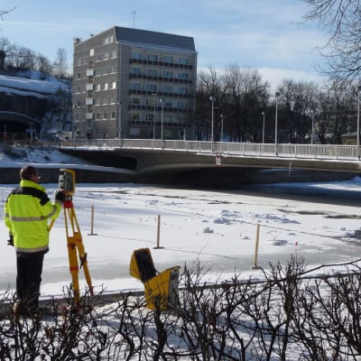 En arbetare med mätinstrument i förgrunden. I bakgrunden den kollapsade Kvarnbron i Åbo.