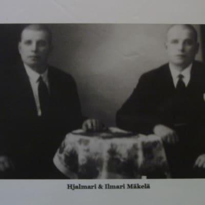 Tvillingbröder Hjalmari och Ilmari Mäkelä sitter vid ett bord på ett svartvit fotografi. 