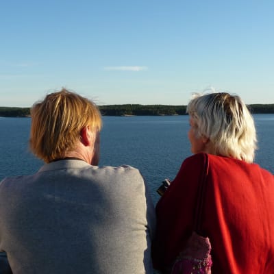 En man i grå tröja och en kvinna i röd tröja står på ett fartygsdäck och tittar ut över en stilla skärgård.