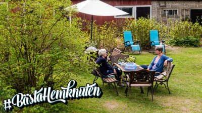 Två kvinnor sitter i en grönskande trädgård, vid ett bord dukat med kaffekoppar.
