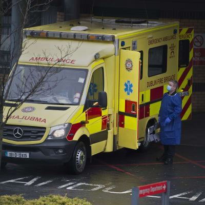 Ambulanssi, jonka vierellä seisoo mies suojavarusteissa.