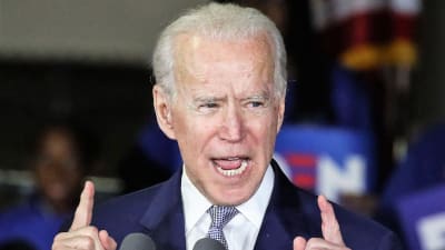 Biden puhuu osoittaen sormillaan ylöspäin. Hänellä on tummansininen puku. Taustalla näkyy Bidenin kannattajia.