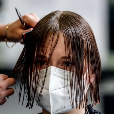 En person med munskydd har vått hår och en frisör klipper personens hår.