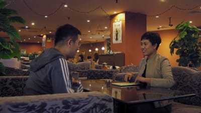 Ett kinesiskt par på blinddejt sitter vid ett cafébord. Kvinnan är prydligt klädd, mannen är klädd i träningsoverall.