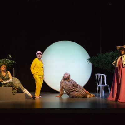 Fyra skådespelare i granna kläder står på en scen med en stor rund lampa i bakgrunden.