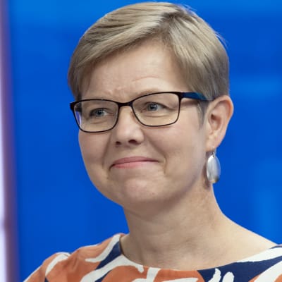 Miljöminister Krista Mikkonen (Gröna) i närbild i en studiomiljö. Hon är i närbild och ler snett mot kameran i en färggrann skjorta. Studioväggen är blå.