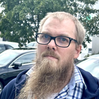 Silmälasipäinen parrakas mies katsoo kameraan.