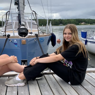 Två unga flickor sitter mot varandra på en brygga framför en stor segelbåt i en hamn. 
