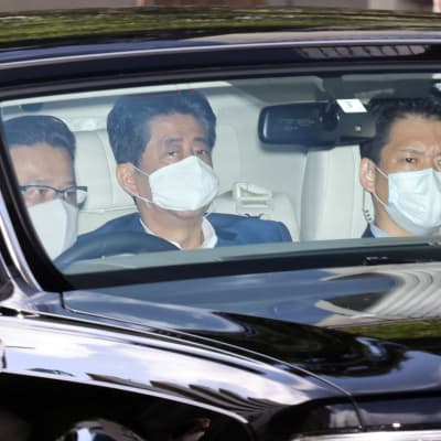 Tre japanska män sitter i en bil och de har munskydd på sig. En av männen är Japans premiärminister Shinzo Abe.