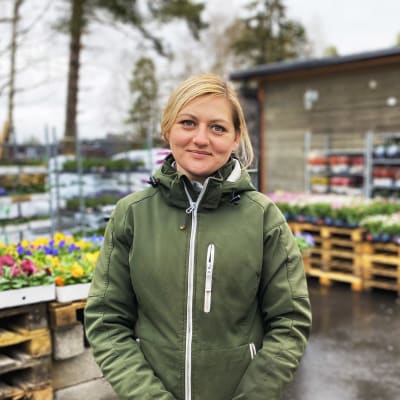 Kvinna med blont hår och grön jacka står framför blommor.