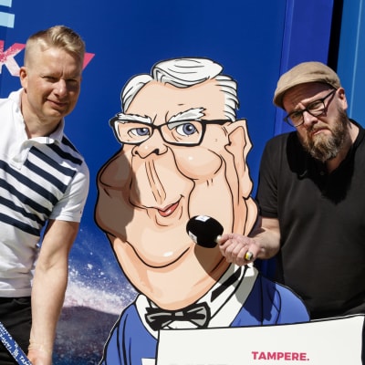 Ylen asiantuntija Juha Juujärvi ja toimittaja Jussi Paasi poseeraavat Kalervo Kummolaa esittävän kuvan vieressä.