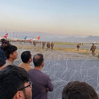 Folk står bakom taggtråd medan amerikanska soldater håller vakt på flygplatsen i Kabul.