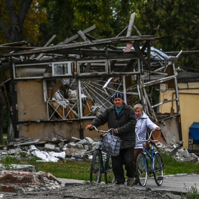 Mies ja nainen taluttaa pyöriä. Taustalla tuhoutunut rakennus.