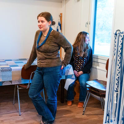 En ung kvinna i brun tröja, jeans och scouthalsduk står och lutar sig mot en stol vid ett bord. I bakgrunden en annan ung kvinna med långt utsläppt hår och rutig tröja som ser ut genom fönstret.