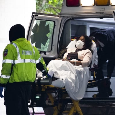 Kasvomaskiin puettu ja paareilla makaava nainen kannetaan ambulanssista sairaalan pihalle