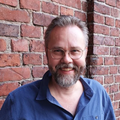 Kuvataiteilija Heikki Marila, harmaapartainen hymyilevä mies pukeutuneena siniseen kauluspaitaan.