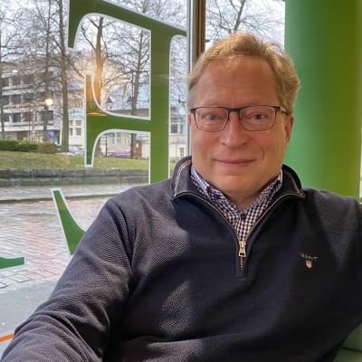 Eläköitynyt apulaiskaupunginjohtaja Juha Isosuo hymyilee kameralle kahvilan pöydän ääressä istuen.