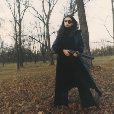 Frigga (Christina Lindberg) står svartklädd med ett vapen i hand i en höstlig skog.