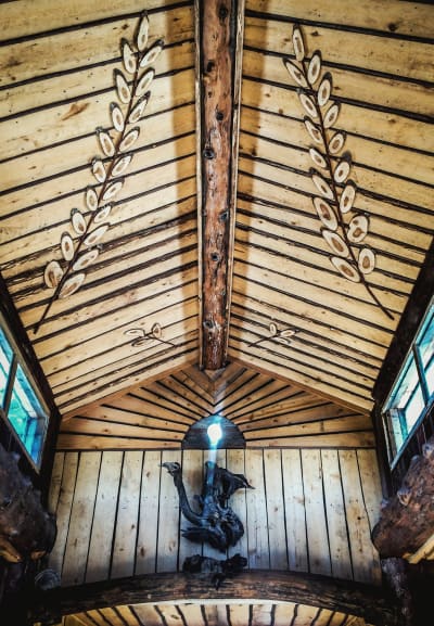 Sisällä Julumetun salissa Kammi-kylässä, päätyseinässä puusta tehty lintumainen koriste, lasinen ikkuna josta loistaa valoa, katossa puusta leikattuja oksamaisia koristeita.