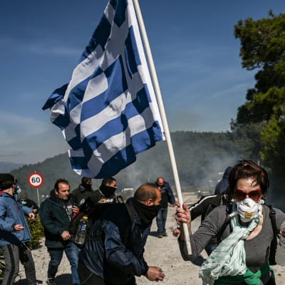 Demonstranter protesterar på ön Lesbos i Grekland. En av demonstranterna håller i en grekisk flagga och de flesta har andningsskydd på sig på grund av polisens tårgas.