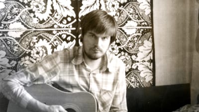 Marit Berndtsons storebror Klaus Berndtson spelar gitarr på en soffa eller säng i ett rum med ett blommigt tyg på väggen bakom honom. Möjligen 1980-talet.